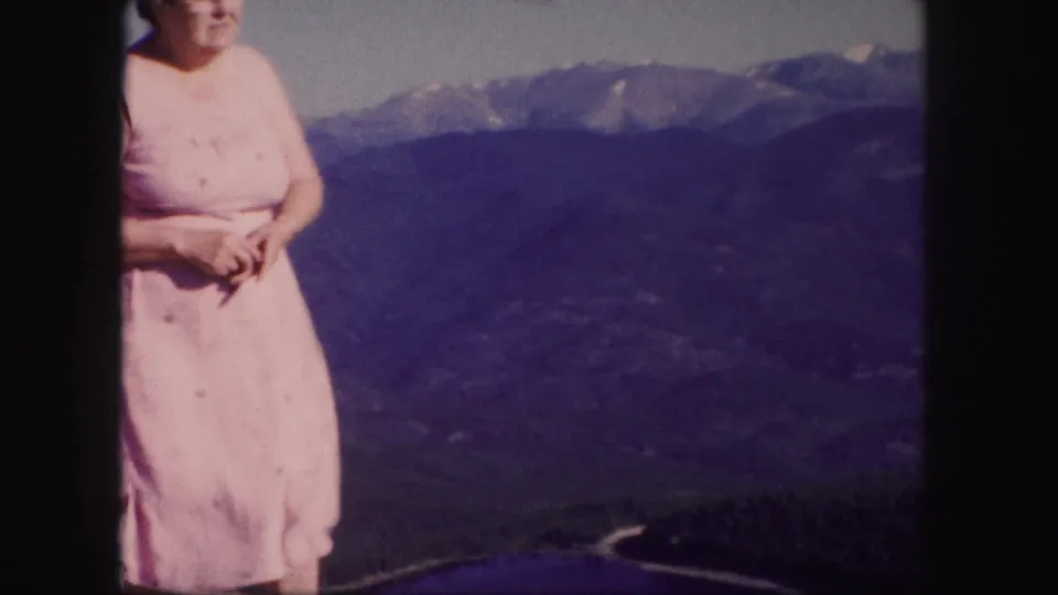 A woman in a pink dress stands near the edge of a Colorado mountain view, with forested slopes and distant peaks behind her.
