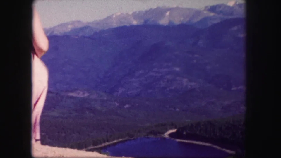 The left edge of the woman remains in frame as the camera favors the mountain slopes and blue lake below.