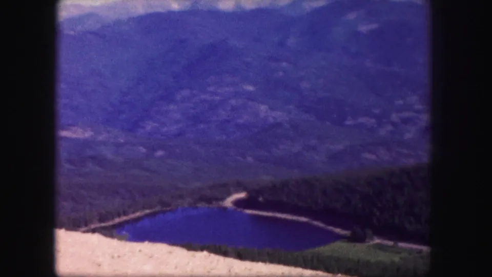 A blue alpine lake sits below dark forested slopes and distant Colorado mountain ridges in a vintage 8mm frame.