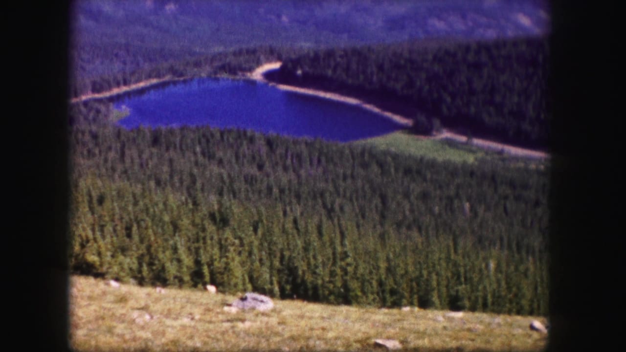 1958 Colorado 8mm view of a blue lake in a forested valley, seen from a high overlook with a pale shoreline road.