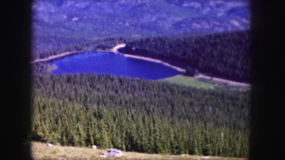 Soft 8mm frame of a Colorado mountain lake in 1958, with forest, a shoreline road, and hills beyond.