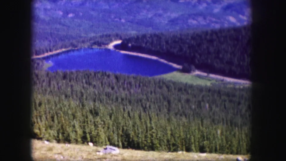 1958 Colorado 8mm view of a blue mountain lake below a wooded slope, with a road or path along the shore.