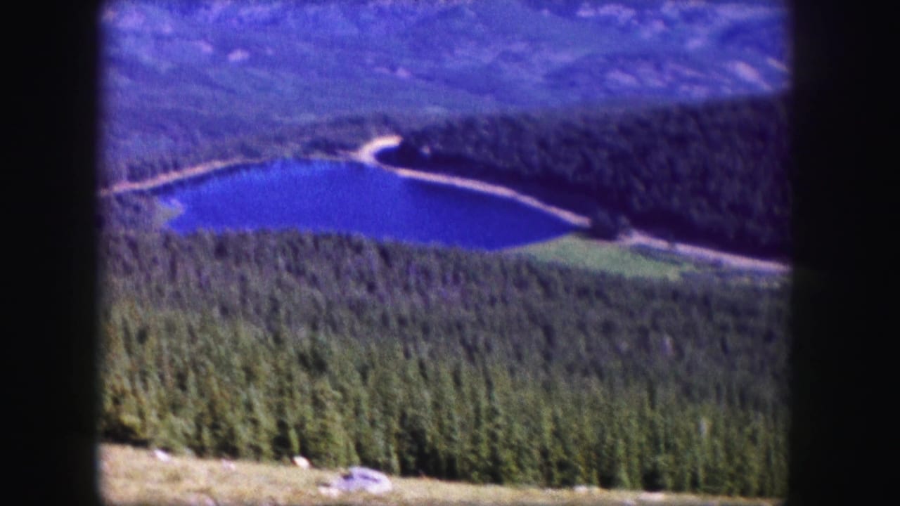 1958 Colorado home-movie frame showing a deep blue alpine lake, surrounding forest, distant mountains, and visible film gate edges.