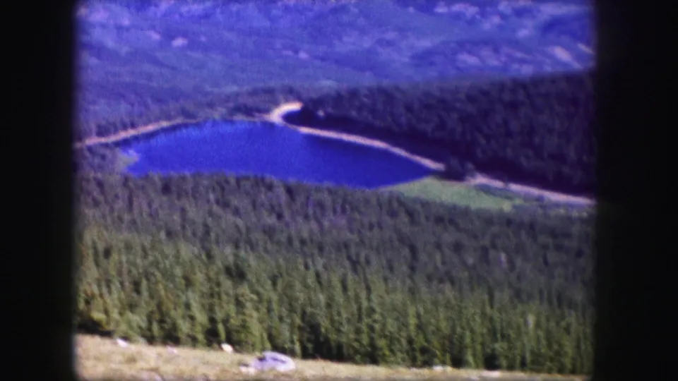 1958 Colorado home-movie frame showing a deep blue alpine lake, surrounding forest, distant mountains, and visible film gate edges.