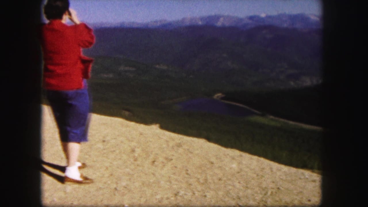 Woman in a red top and blue shorts stands on a mountain overlook facing distant peaks.