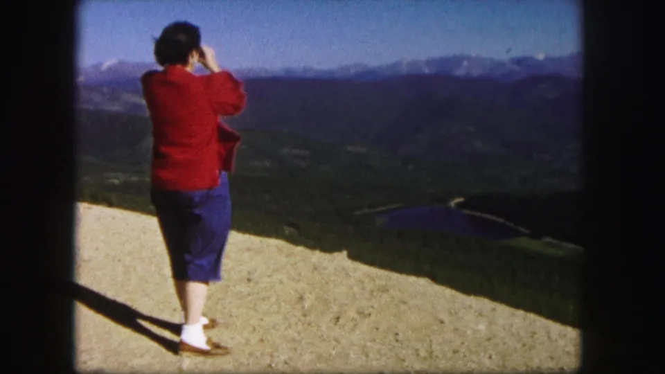 1958 Colorado home-movie frame showing a woman from behind at a high overlook, with forested slopes, a small lake, and distant peaks ahead.