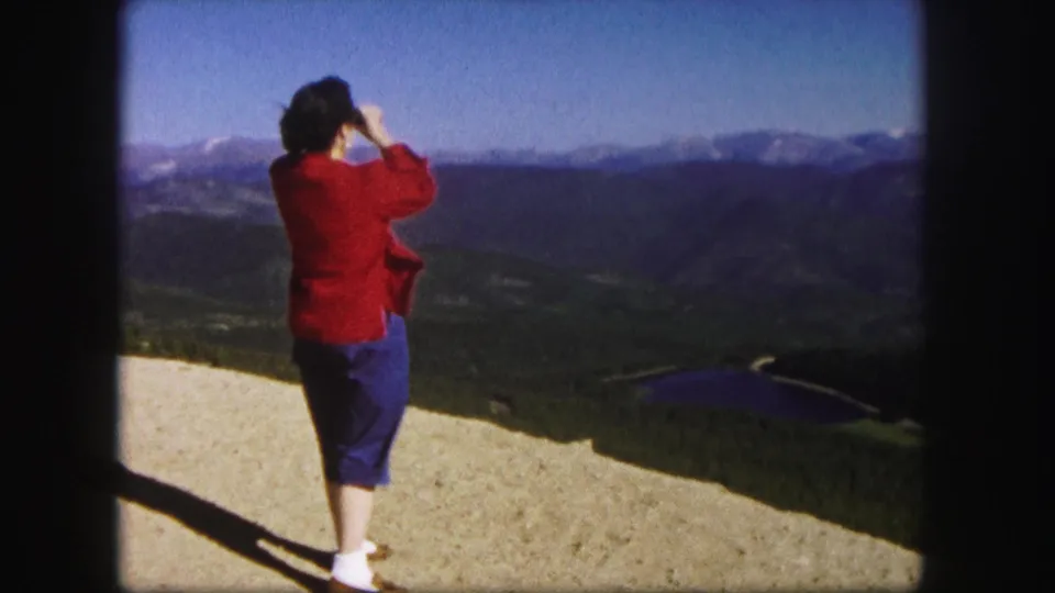 1958 Colorado home-movie frame of a woman viewing or photographing a mountain valley with a dark lake and distant snow-touched ridges.