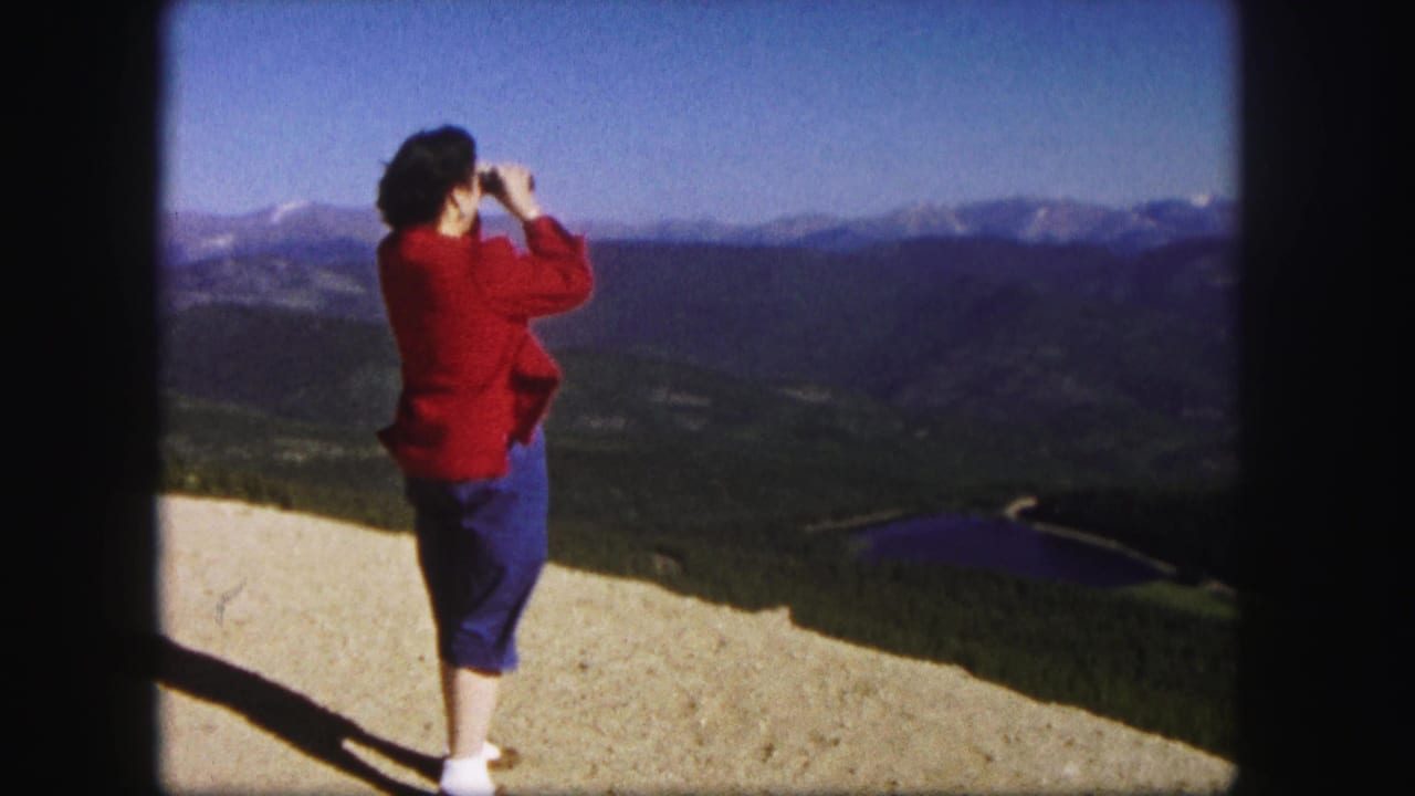 1958 Colorado 8mm frame of a woman in a red sweater looking through binoculars across mountain ridges and a distant lake.