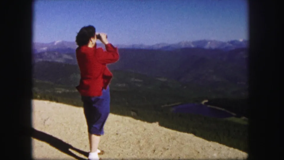 1958 Colorado 8mm frame of a woman in a red sweater looking through binoculars across mountain ridges and a distant lake.