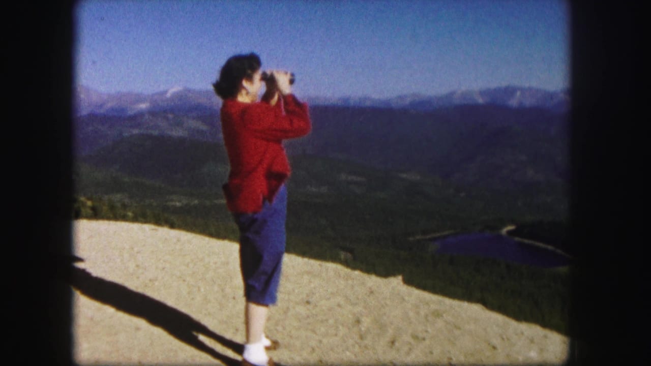 Woman with binoculars standing beside a mountain overlook landscape.