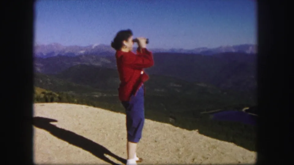 A closer 1958 Colorado home-movie view shows the woman using binoculars, with ridges and a lake stretching behind her.