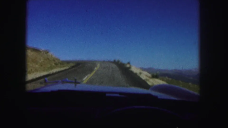 Passenger-view 8mm frame from a car on a Colorado mountain road, with the hood visible beneath a clear sky and roadside slope.