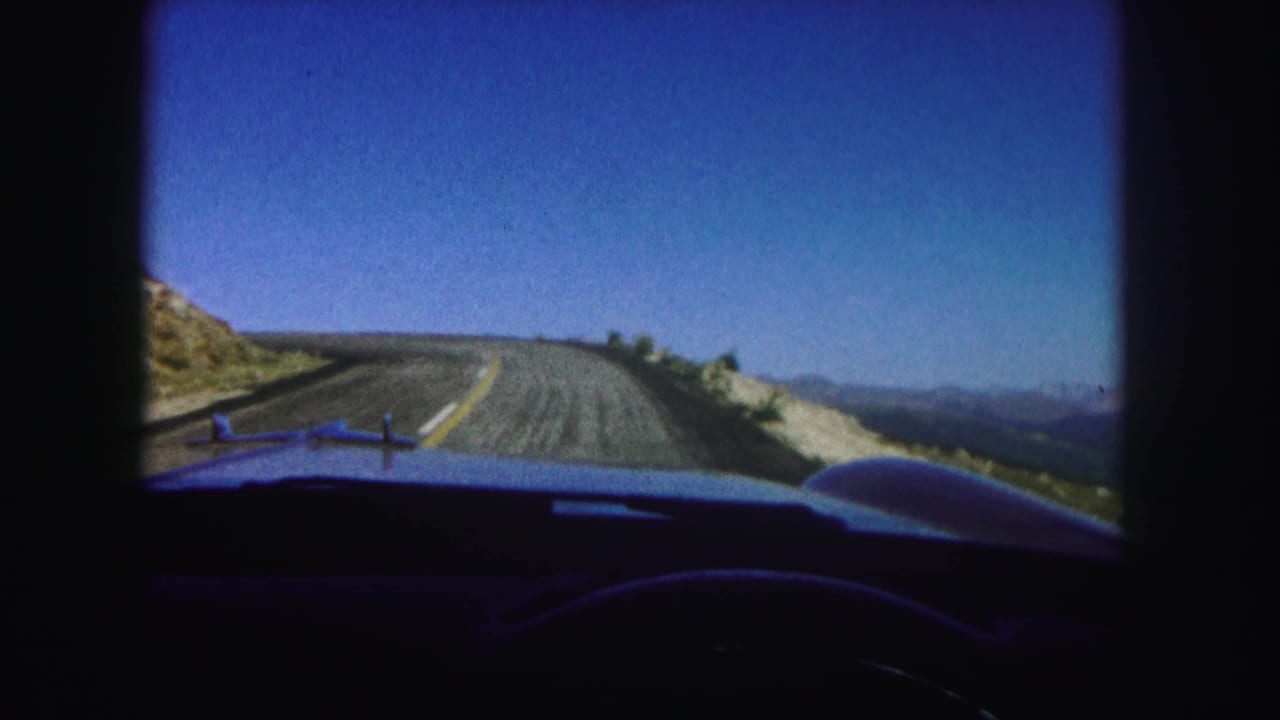 1958 Colorado home-movie frame looking over a car hood at a winding mountain road with dry slopes and distant ridges.