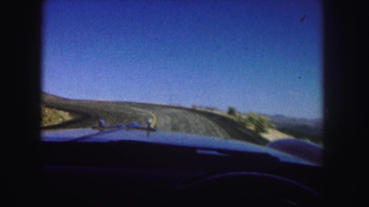 Car windshield view of a road bending across dry mountain terrain.
