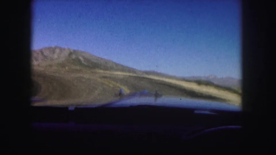 1958 Colorado 8mm frame from inside a car, showing the road ahead, rocky hillsides, and distant mountain forms.
