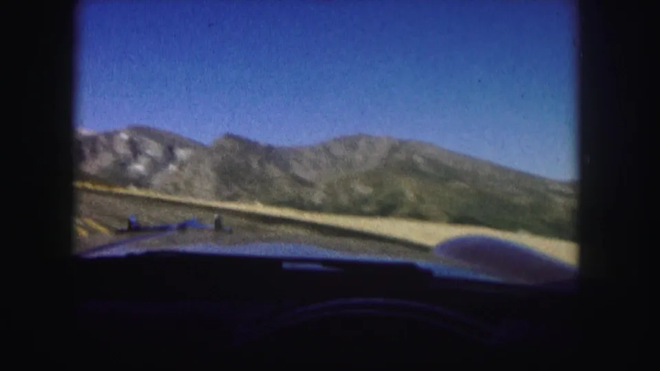 1958 Colorado windshield POV with the car hood in front, a mountain road ahead, and rocky ridgelines under a clear sky.
