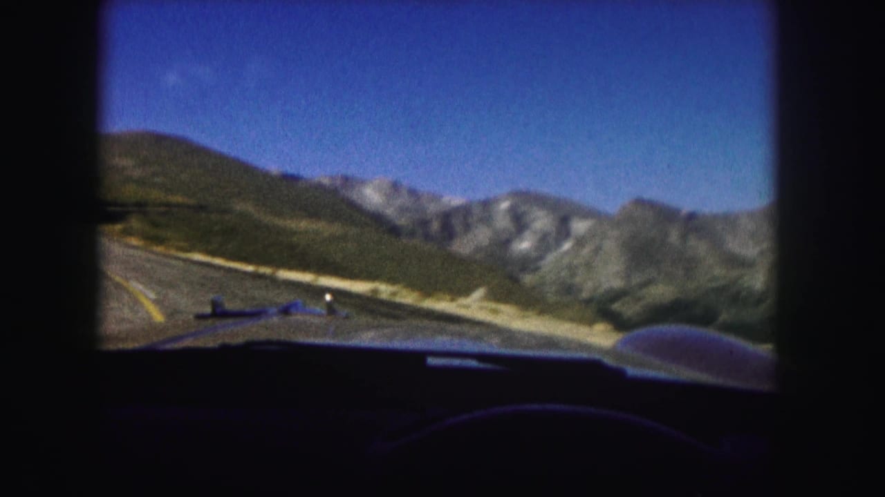 1958 Colorado 8mm frame looking from inside a car along a paved mountain road toward rugged peaks under a blue sky.