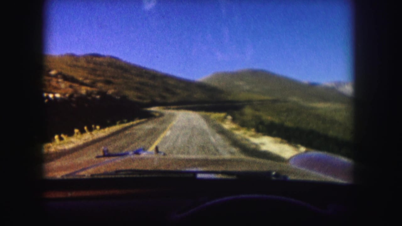 Mountain road curving through open slopes with car hood and dashboard visible at the bottom.