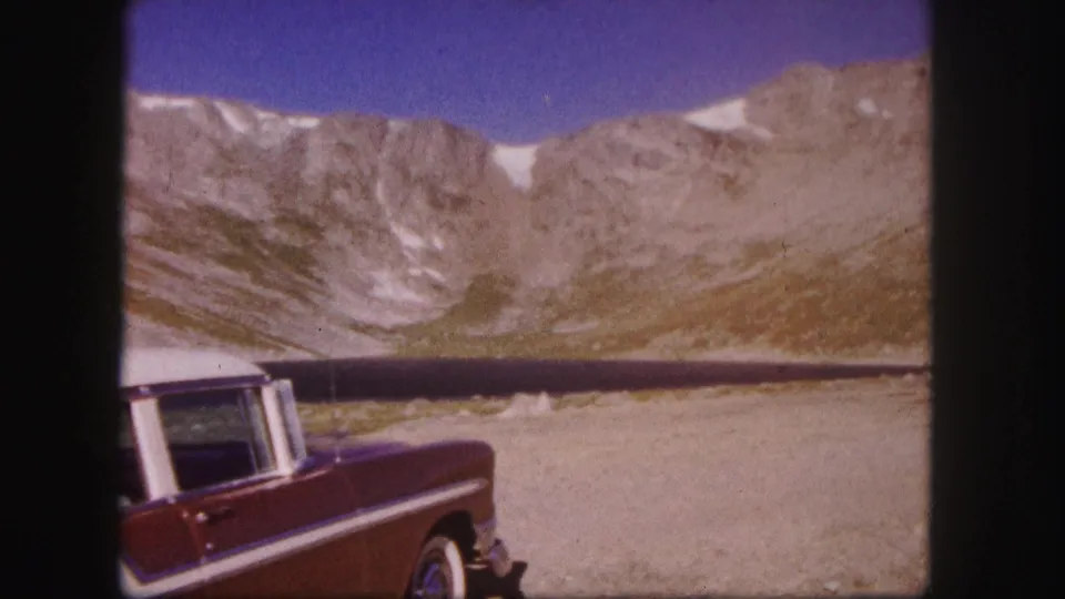 A red mid-century car appears parked near water in the Colorado mountains, with rocky slopes and patches of snow in the background.
