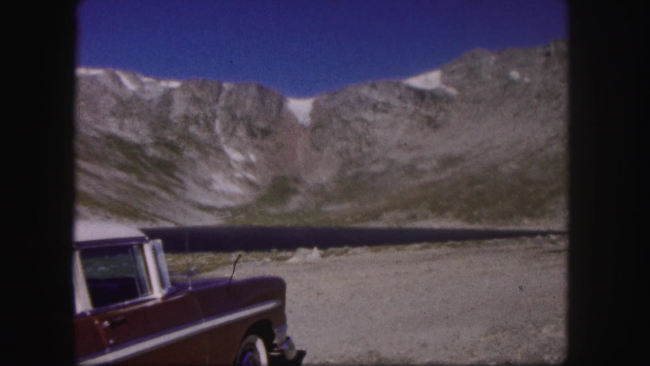 Red car parked beside a dark mountain lake below steep cliffs.