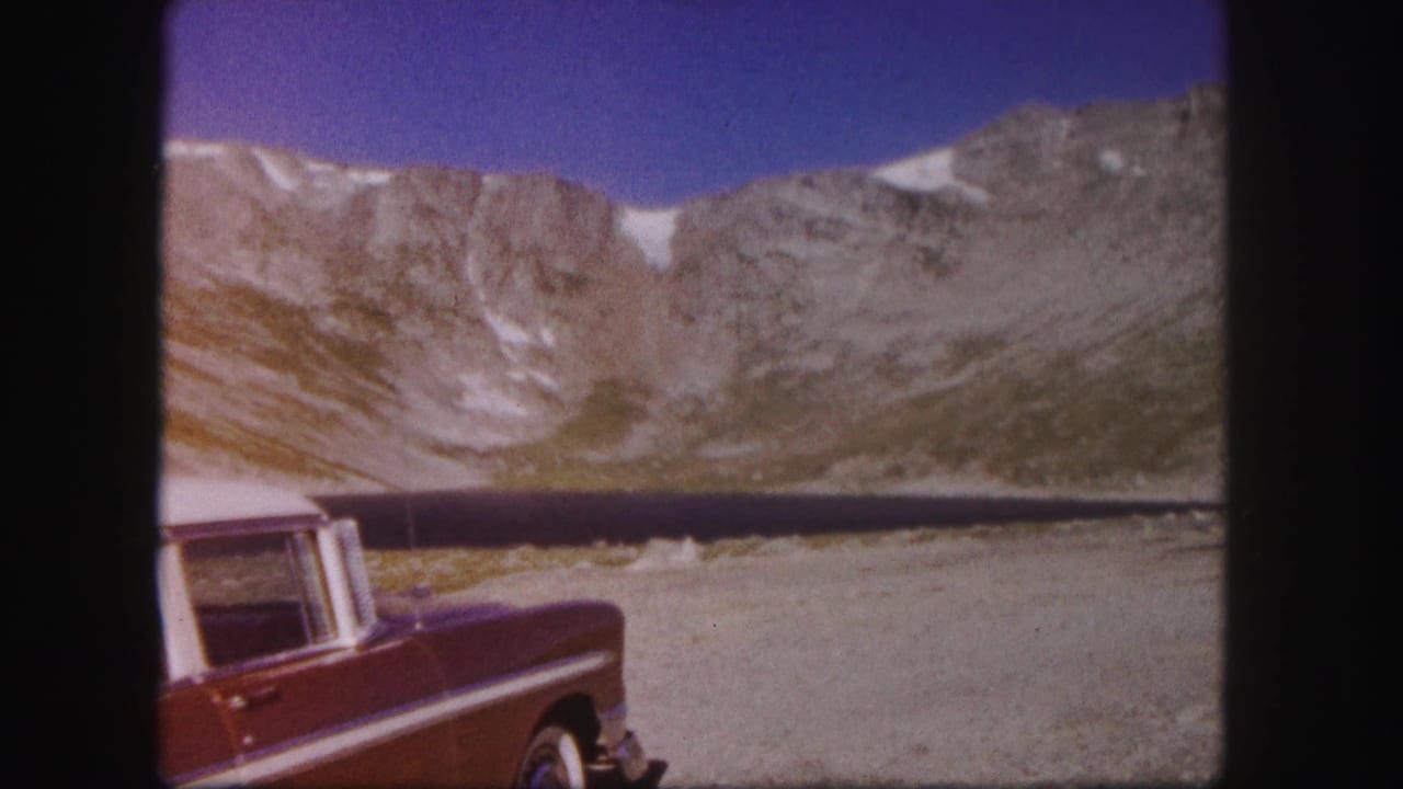 1958 Colorado home-movie frame with a red car in the foreground, a still alpine lake, and sheer rocky slopes under blue sky.