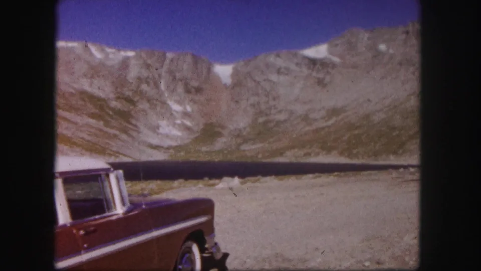 Colorado 1958 8mm frame showing the rear side of a red car near a calm mountain lake backed by pale rocky cliffs.