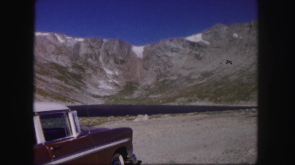 1958 Colorado 8mm frame of a red car partly cropped at a lakeside overlook, with dark water, cliffs, snow traces, and blue sky.