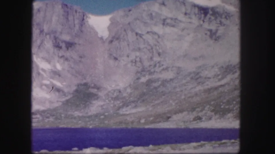 Colorado 8mm home-movie frame from 1958 showing a still alpine lake beneath pale, rugged mountain walls.