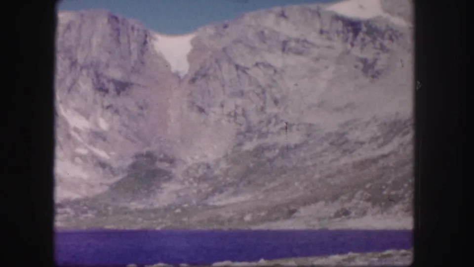 1958 Colorado mountain landscape on 8mm film, with blue lake water below a rugged slope and a small white snowfield.
