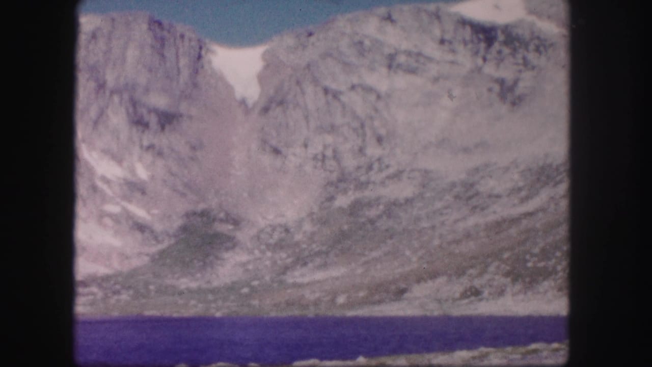 1958 Colorado home-movie frame showing rocky mountain slopes with snow patches above a blue-purple lake.
