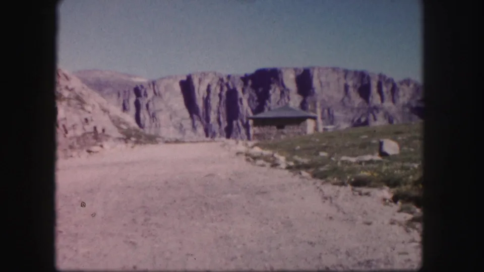 1958 Colorado home-movie view down an unpaved road toward a small stone shelter set below a pale rocky cliff.