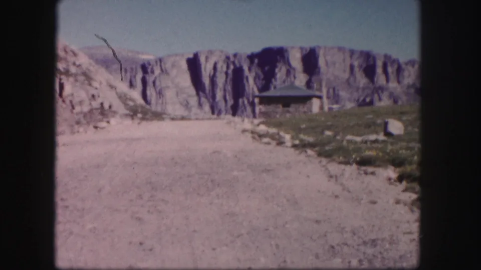 1958 Colorado 8mm frame with a dirt road, sparse alpine ground, and a small shelter below a rugged cliff face.