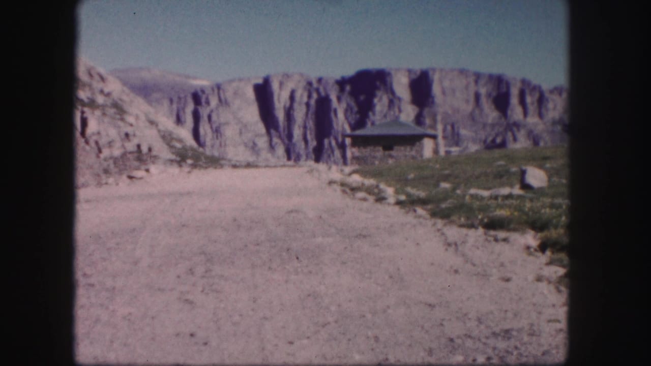1958 Colorado home-movie frame showing a rustic roadside shelter below pale cliffs and an unpaved mountain road.