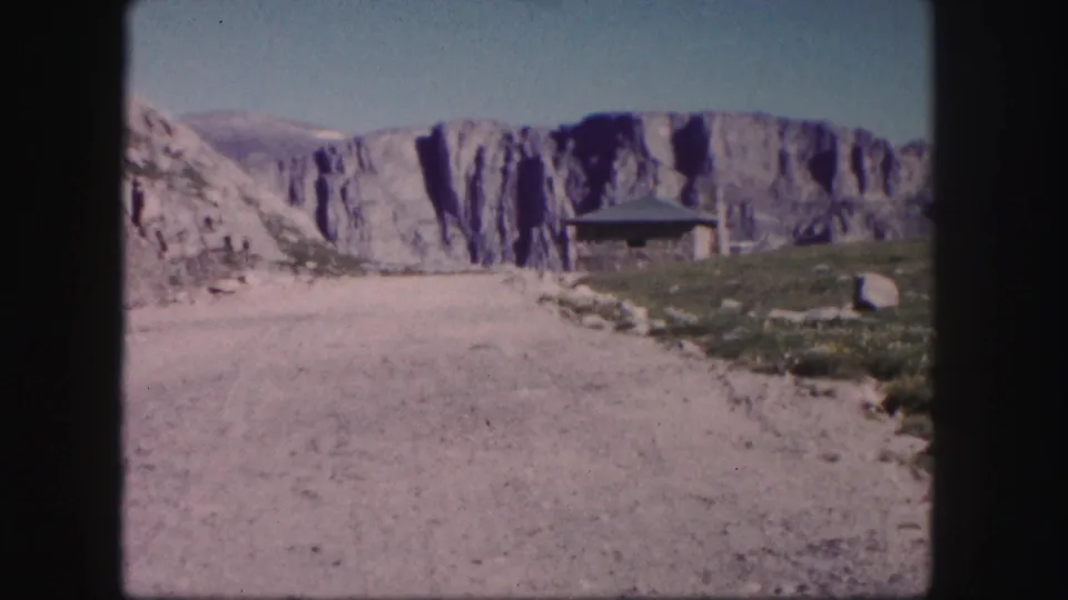 1958 Colorado home-movie frame showing a rustic roadside shelter below pale cliffs and an unpaved mountain road.