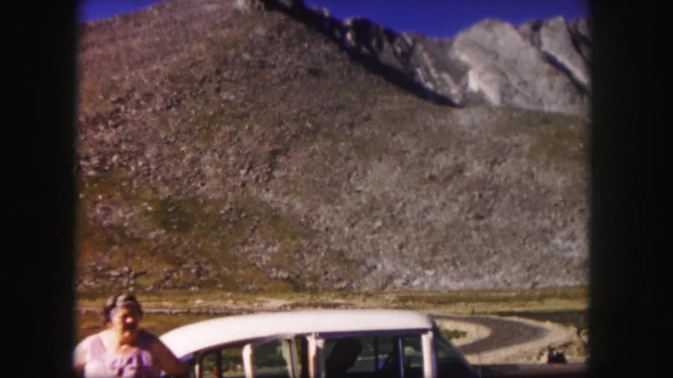 1958 Colorado home-movie frame showing a woman beside a two-tone station wagon below steep rocky slopes.
