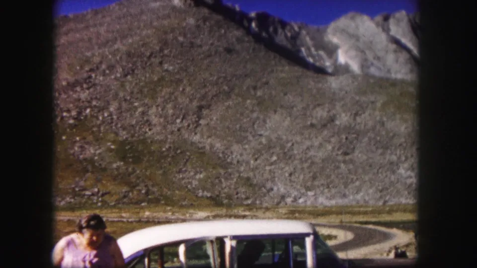 1958 Colorado frame with a woman beside a parked station wagon, a curving road, and high rocky terrain in the background.