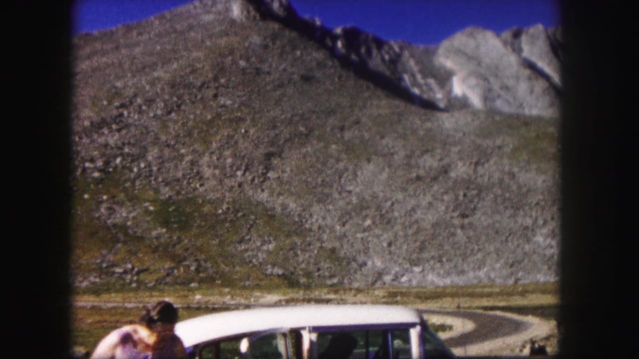 Woman beside a white car below a rocky mountain slope and blue sky.