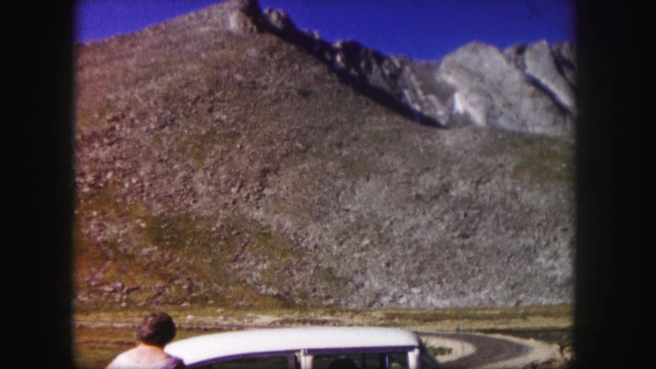 1958 Colorado home-movie frame of a woman by a white car, with a curving mountain road and rocky slope in the background.