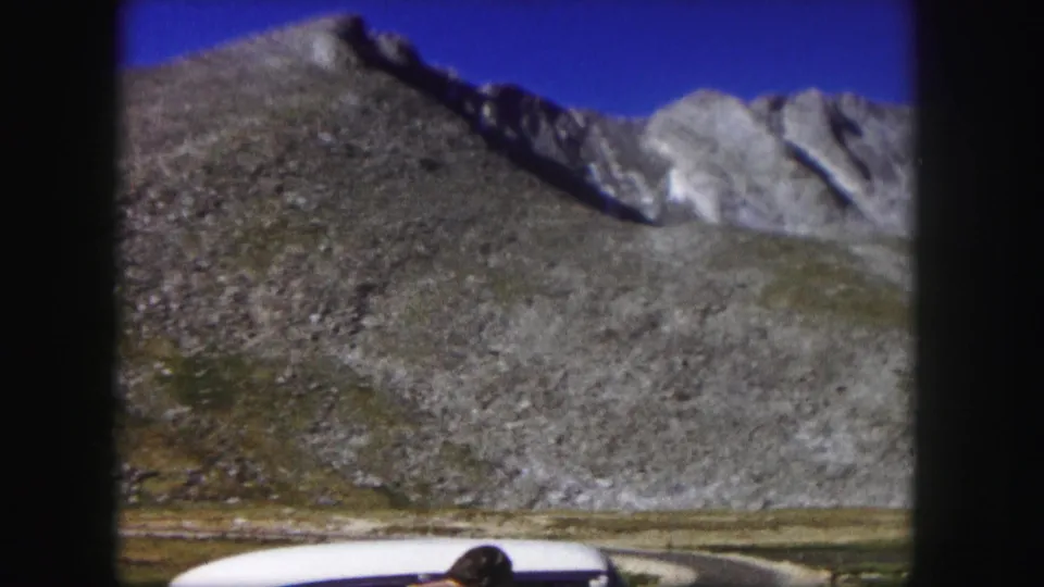 1958 Colorado 8mm frame with the white car partly visible below a broad rocky mountain slope and blue sky.