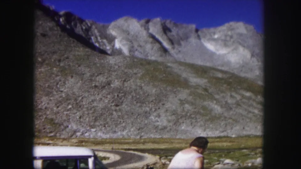 1958 Colorado home-movie frame showing the woman partly at lower right near the white car, with rugged mountain terrain behind.