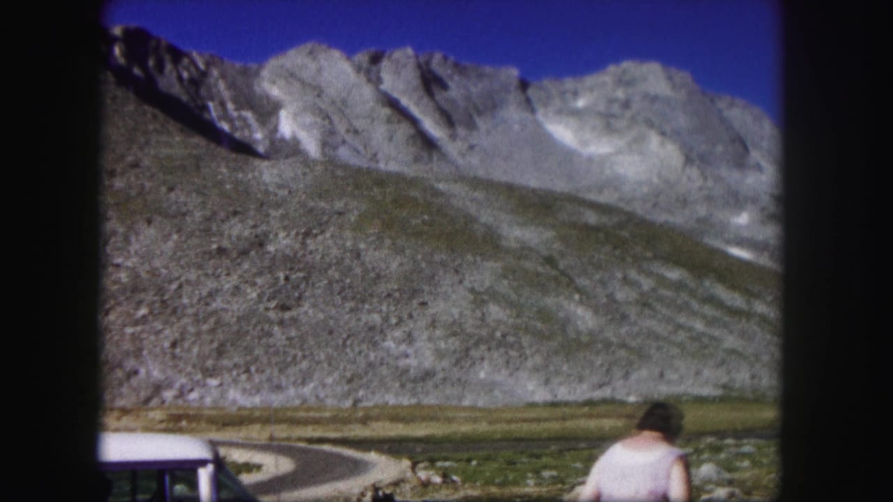 1958 Colorado 8mm frame showing a woman near a rocky roadside below gray mountain peaks and clear blue sky.