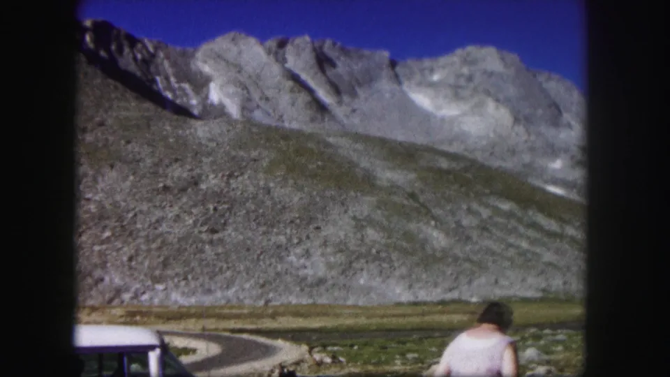 1958 Colorado 8mm frame showing a woman near a rocky roadside below gray mountain peaks and clear blue sky.