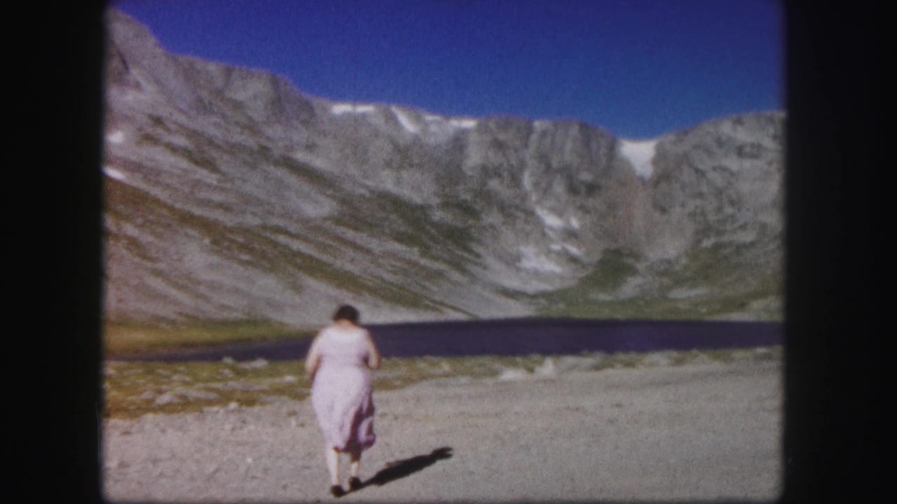 1958 Colorado home-movie frame with a woman walking away from camera toward a dark alpine lake and rugged peaks.