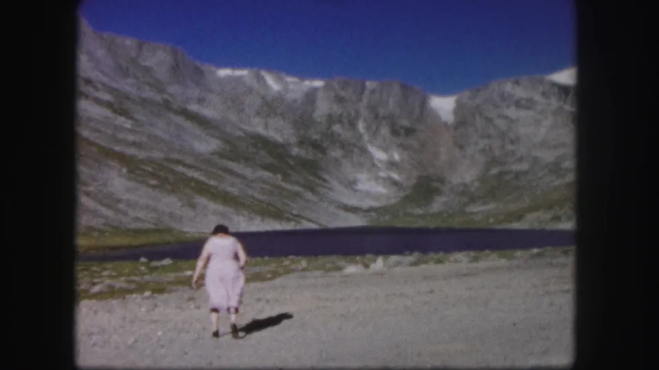 1958 Colorado 8mm frame showing the woman from behind on a rough path beside a mountain lake.