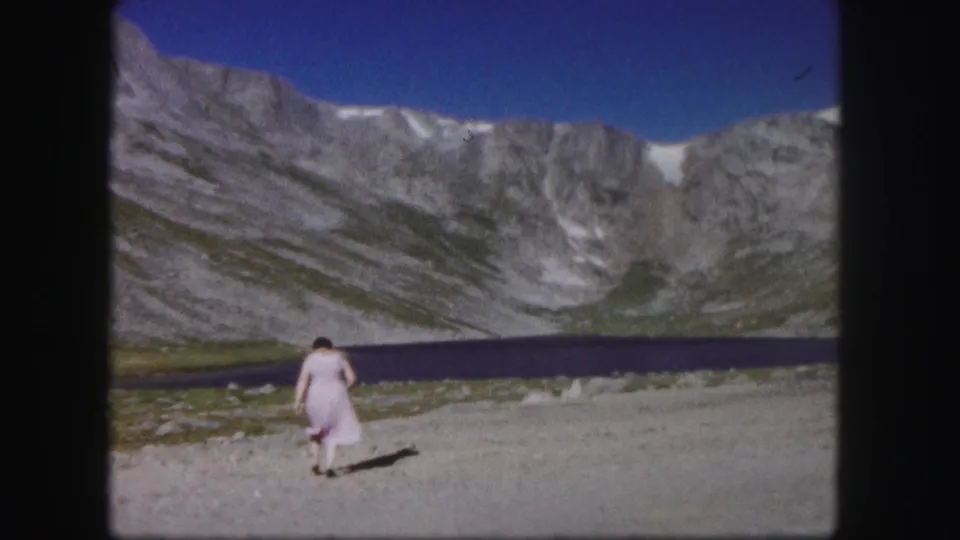 1958 Colorado home-movie frame with the woman smaller in the scene, walking across rocky lakeside ground under steep mountains.
