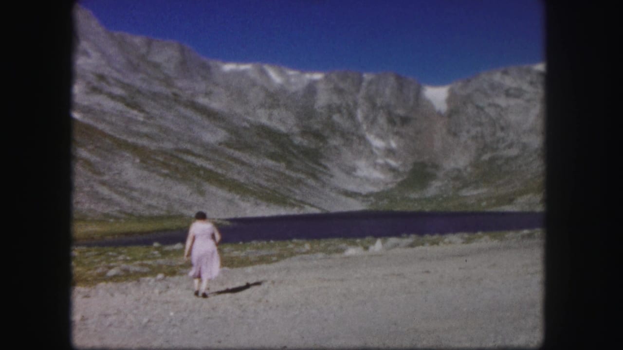 In 1958 Colorado home-movie footage, a woman in a pale dress walks away from the camera toward a deep blue alpine lake below steep mountains.