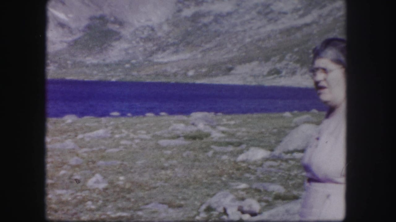A woman in a light dress appears at the edge of the frame beside a blue alpine lake and rocky Colorado mountain landscape.