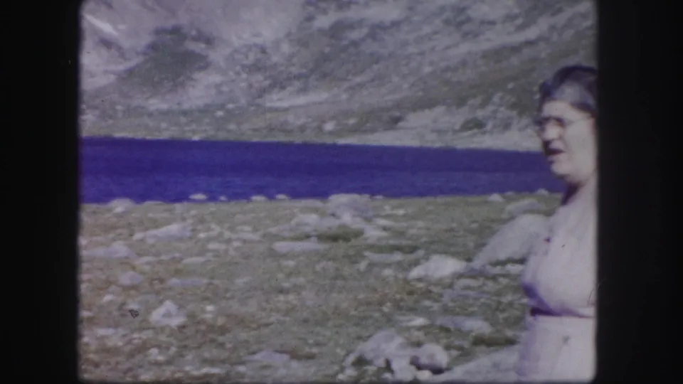 A woman in a light dress appears at the edge of the frame beside a blue alpine lake and rocky Colorado mountain landscape.
