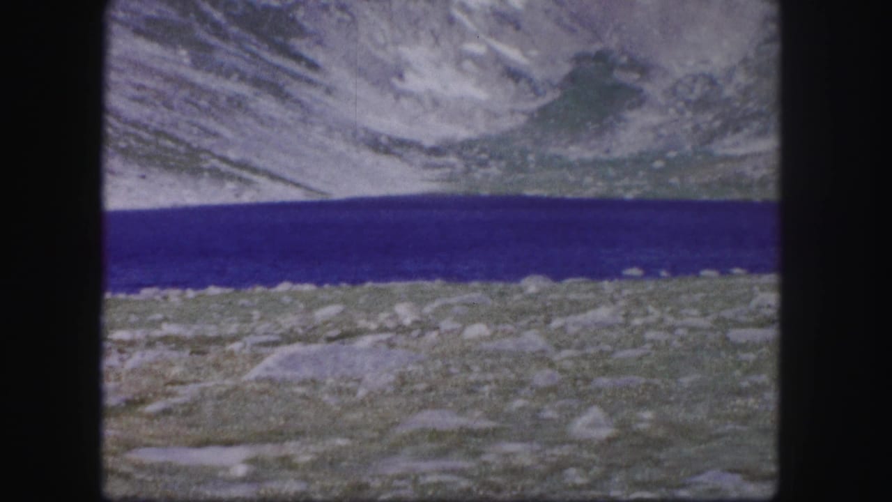 1958 Colorado 8mm frame showing a saturated blue alpine lake, rocky foreground, and pale mountain slopes in soft home-movie focus.