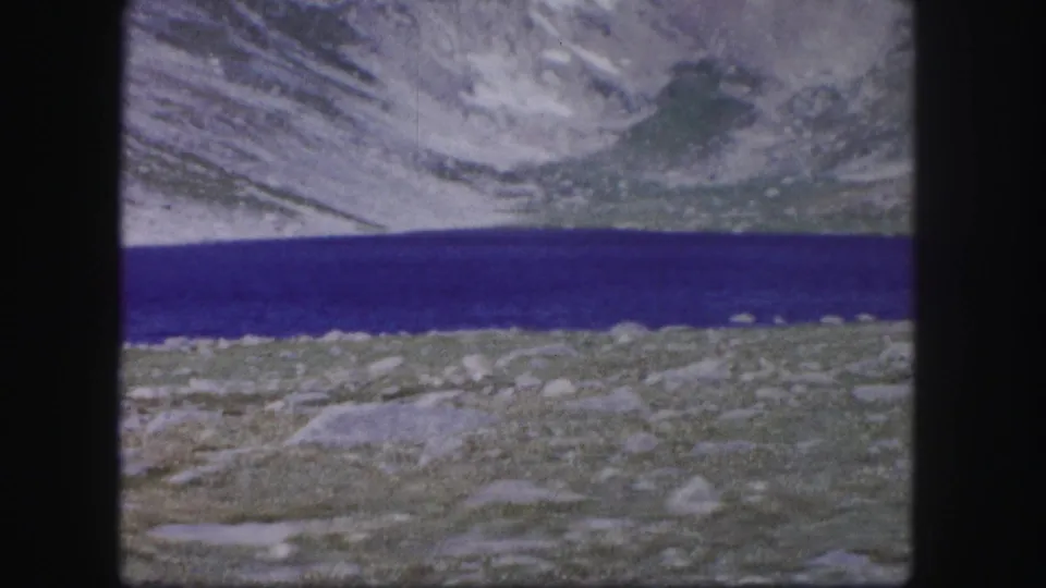 1958 Colorado 8mm frame showing a saturated blue alpine lake, rocky foreground, and pale mountain slopes in soft home-movie focus.